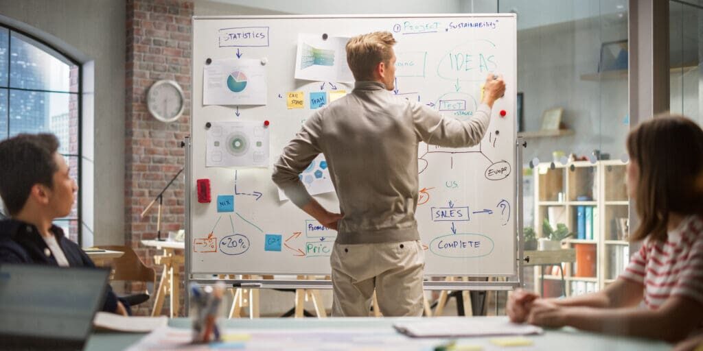 Group Businesspeople have Meeting in Office Conference Room. Crisis Manager Using Charts on Whiteboard, Showing Plan how to Save Corporate Strategy After Bad Quarter. Back View.